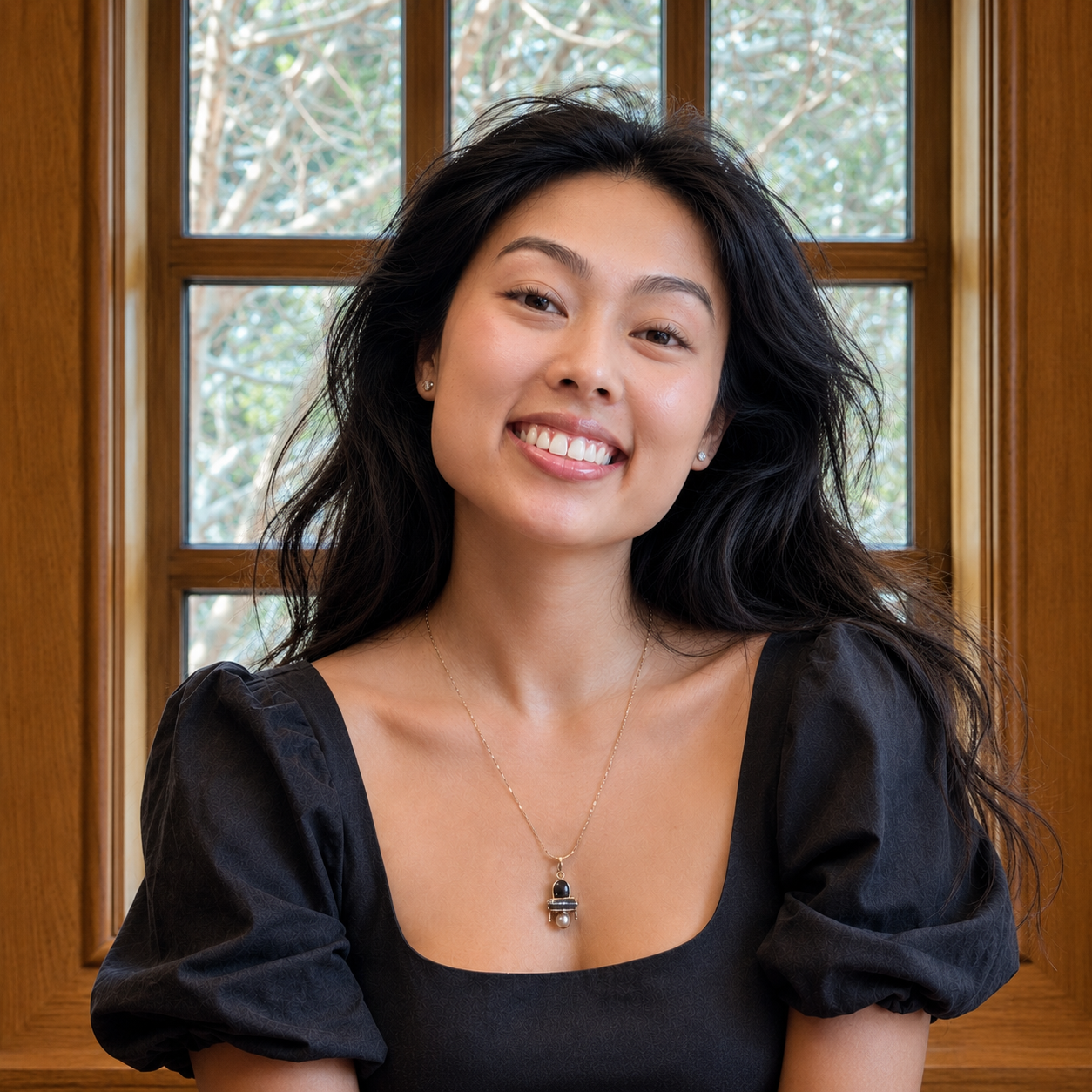 A smiling young woman with long dark hair is shown from the chest up, wearing a black puff-sleeve top and a delicate necklace with a small dark pendant, standing in front of a warm wood-paneled window. The founder of savath wellness.